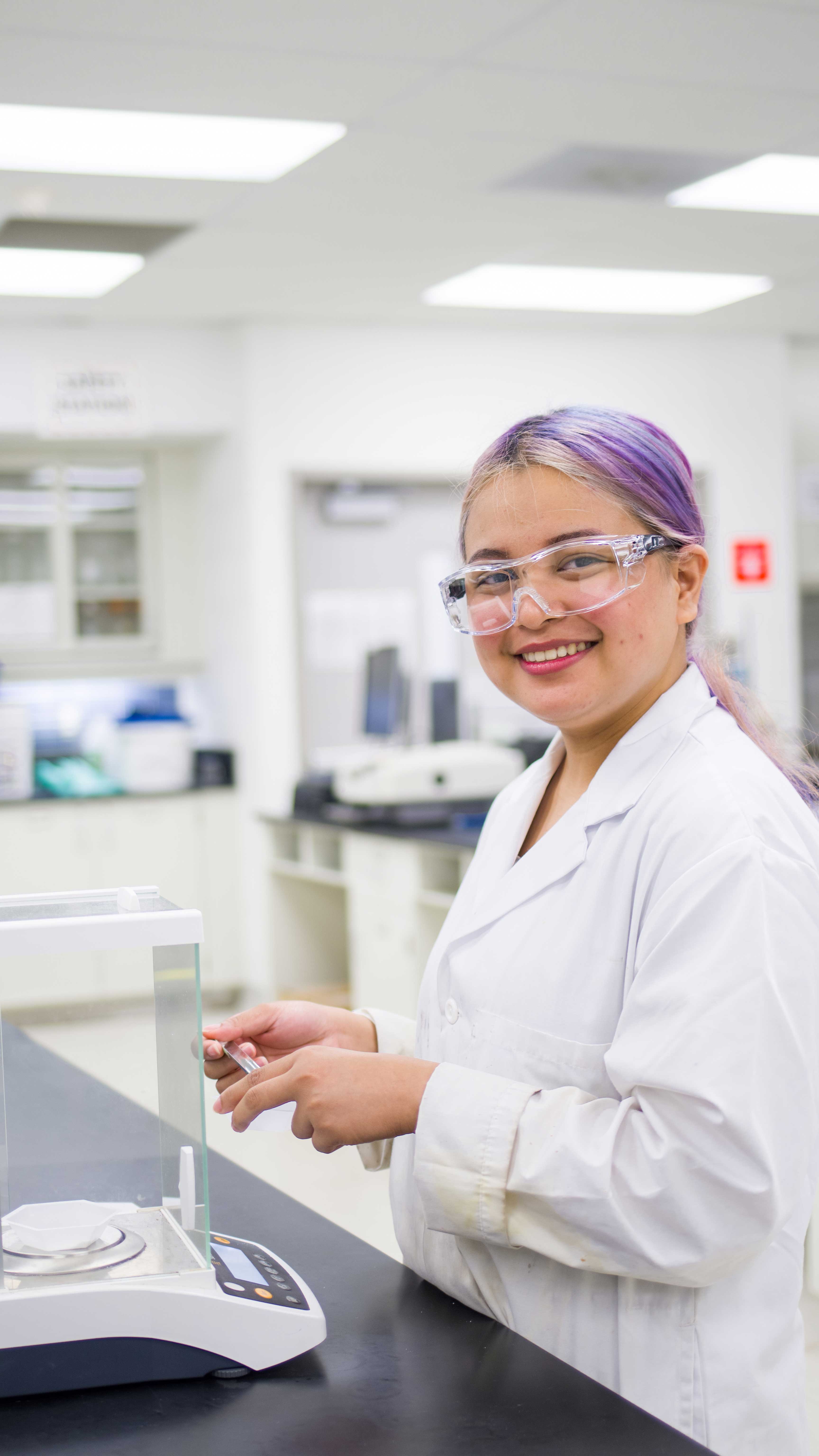 A smiling student with purple hear wearing safety glasses and a white lab coat is looking at the camera while holding a scoopula in front of an analytical balance